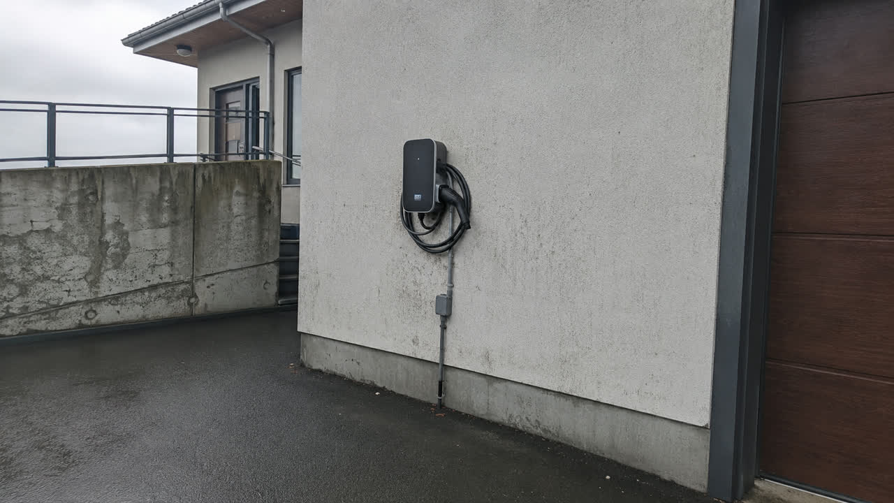 Hleðslustöð fyrir rafbíl fest á múraðan útvegg við bílskúrshurð, lögn í röri niður vegginn, blautt malbik og skýjað veður.